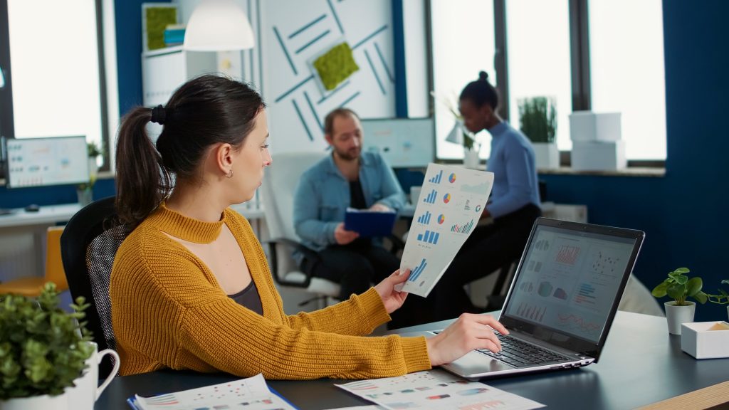 Woman comparing statistics with sales charts on laptop and taking a break to drink coffee or tea